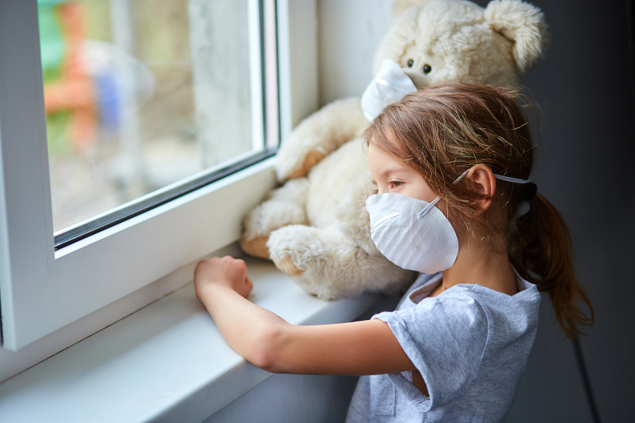 Little girl holding and hugging teddy bear in mask near the window. Przygnębiona mama w obliczu koronawirusa